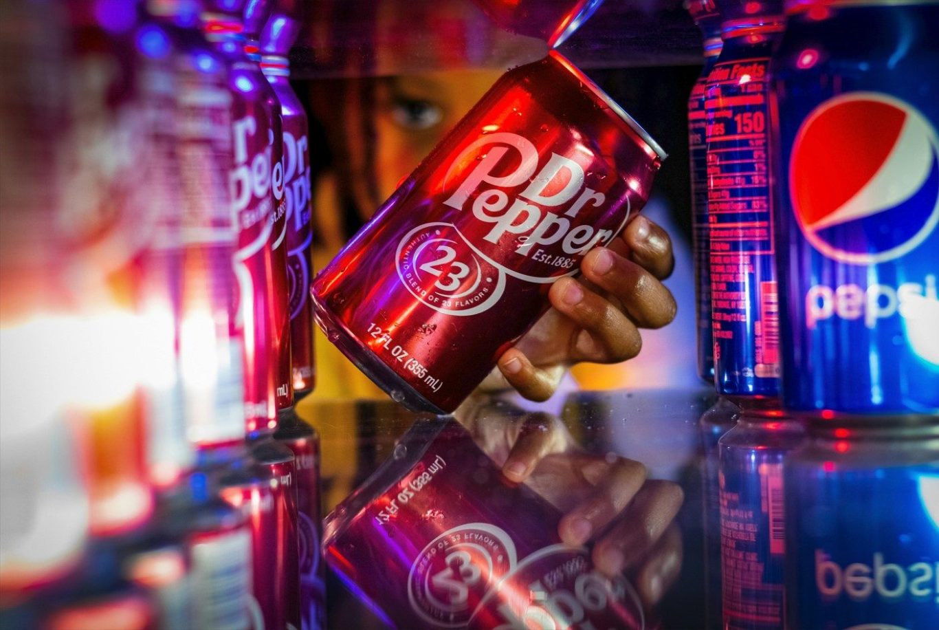 Dosen Eine Hand hält eine Dose Dr. Pepper zwischen leuchtenden Flaschen von Pepsi als Beispiel für ein gutes Brand Naming.